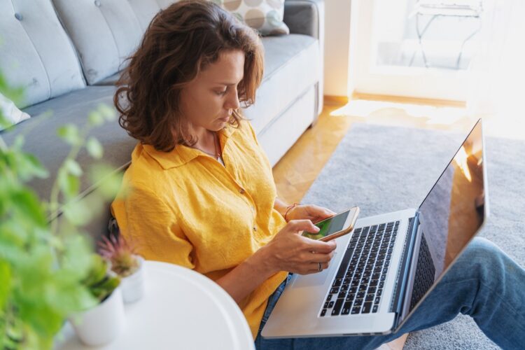 A beautiful young woman in a yellow shirt sits on the floor on a carpet with a laptop.