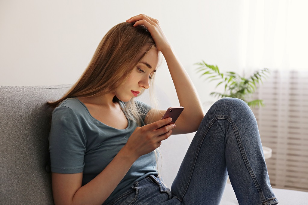 Beautiful young woman with depressed expression sitting on couch holding her phone. 