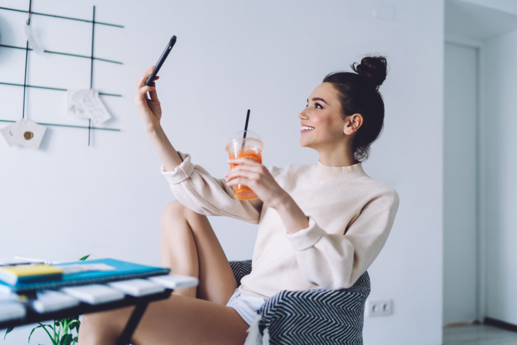 Smiling woman taking selfie with smartphone while sitting in armchair at home