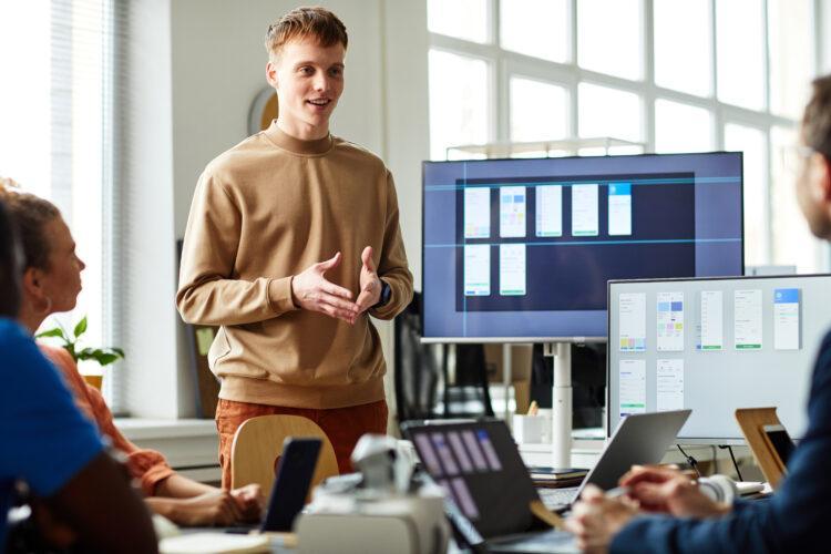 Young IT specialist presenting new project on monitor to his colleagues during meeting at office