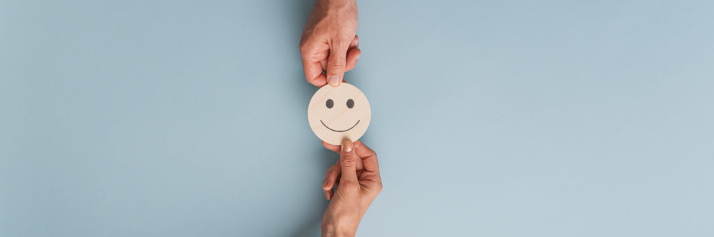 Male and female hands holding wooden cut circle with smiling face on it. Over blue background with copy space.