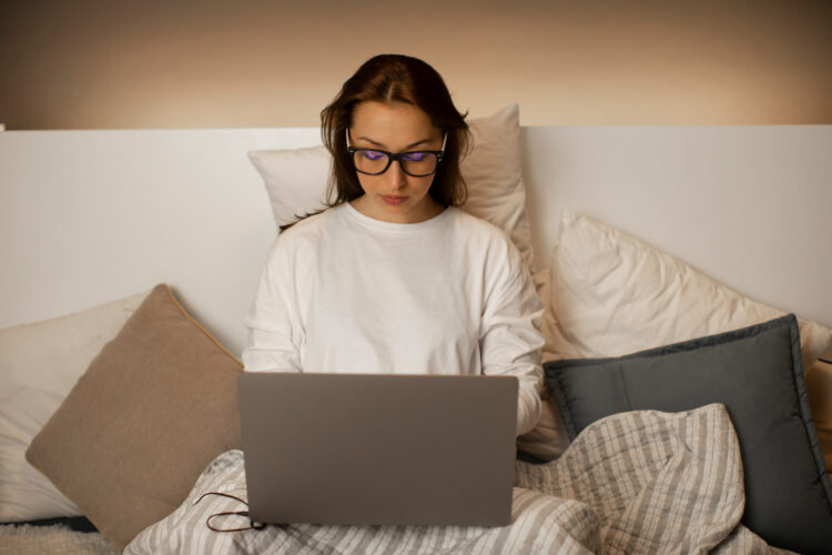 Beautiful woman in glasses sits in bed with laptop, works at home