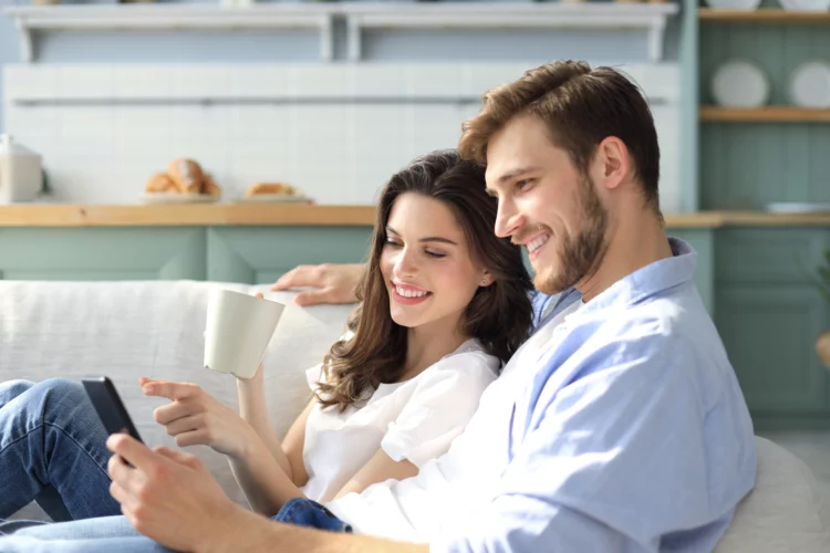 Young couple watching online content in a smart phone sitting on a sofa at home in the living room.