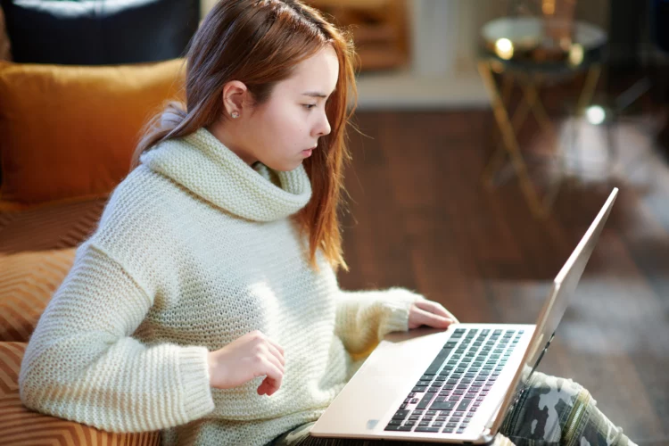 Modern young woman using website while sitting near couch