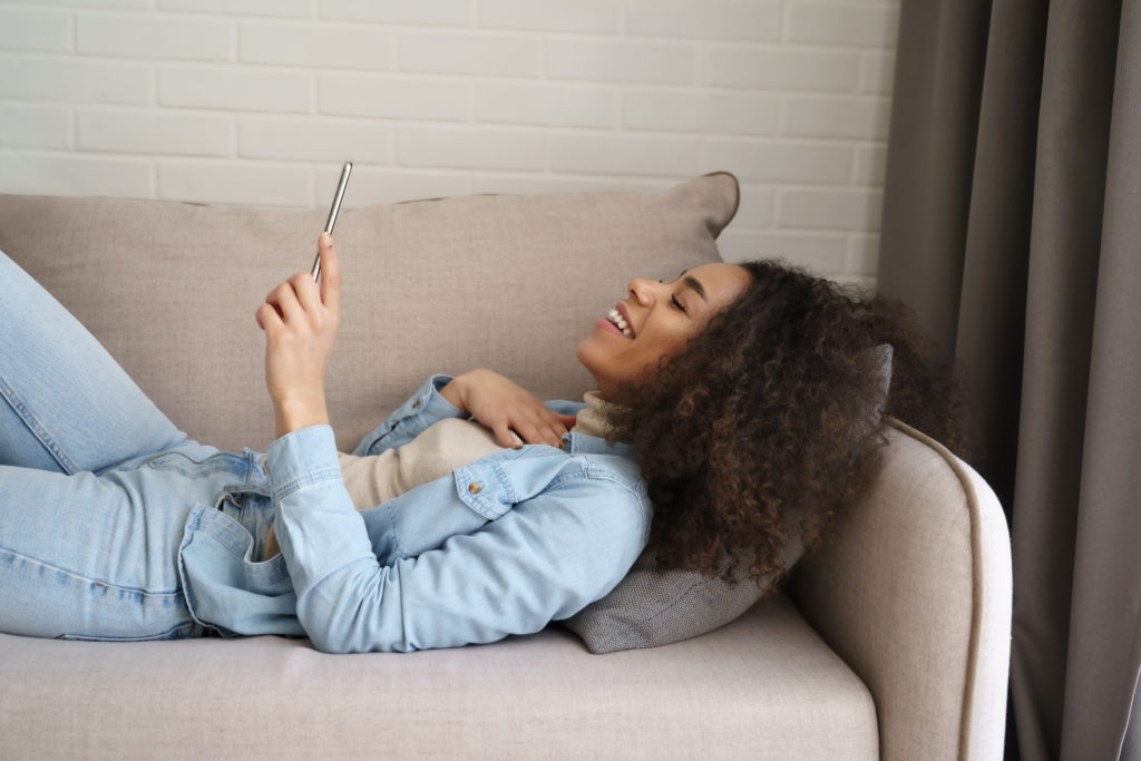 Happy young woman on a sofa laughing while using her phone.