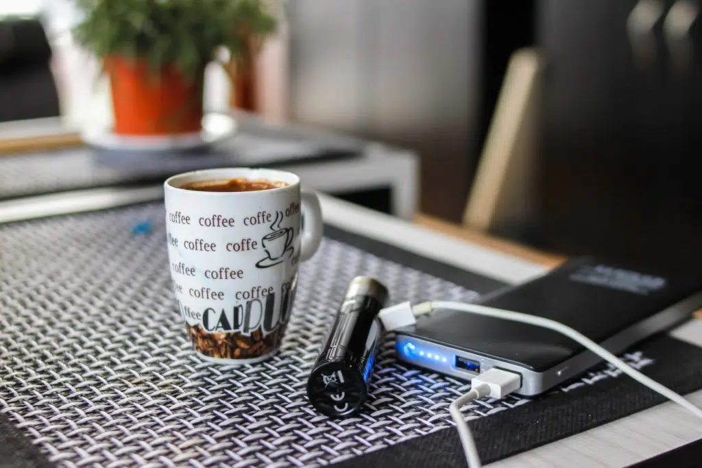 E-cigarette connected to a powerbank beside a cup of coffee.