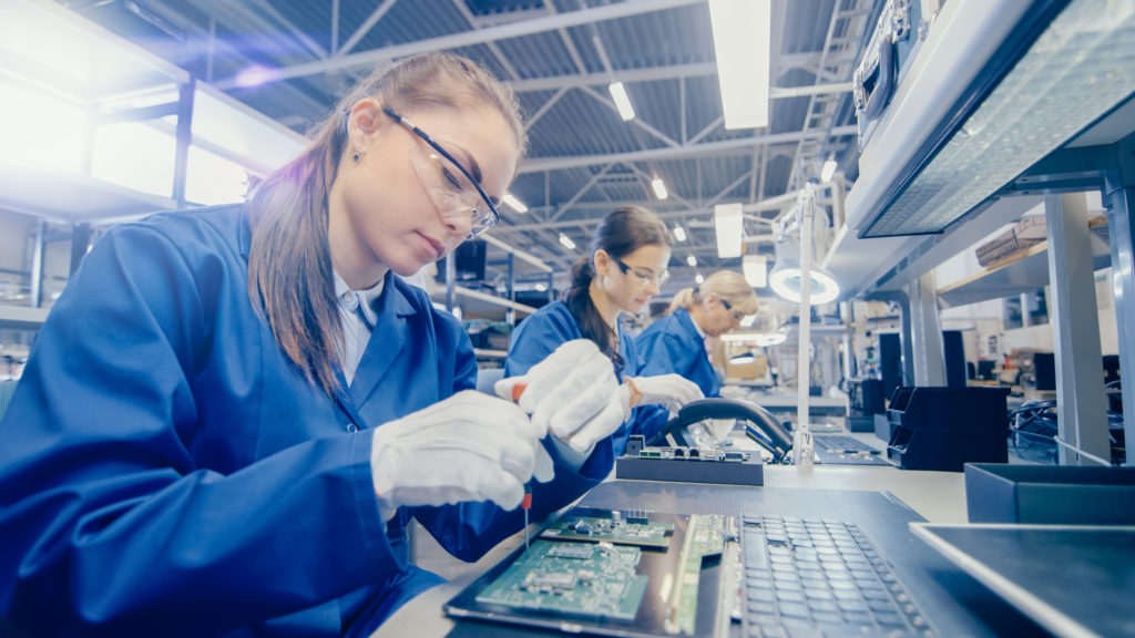 Female Electronics Factory Worker in Blue Work Coat and Protective Glasses is Assembling Laptop's Motherboard with a Screwdriver. High Tech Factory Facility with Multiple Employees.