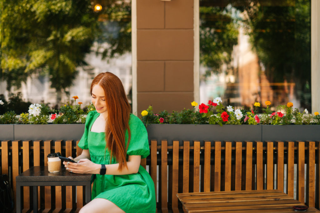 Attractive smiling young woman using smartphone, sitting at table with coffee cup at an outdoor café.