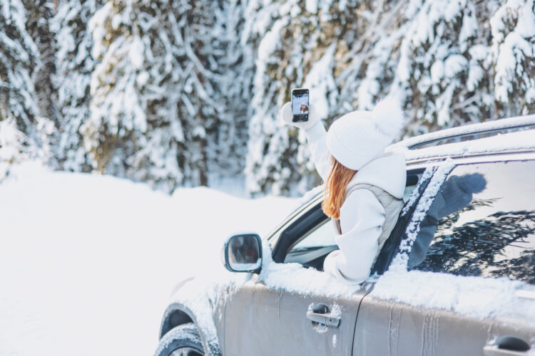 Young woman traveling looking out of car window and taking selfie on smartphone in winter snowy forest.
