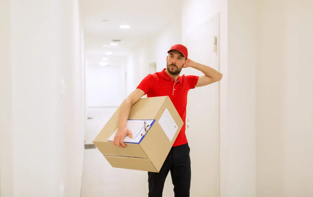 Delivery man with box and clipboard in corridor looking puzzled.
