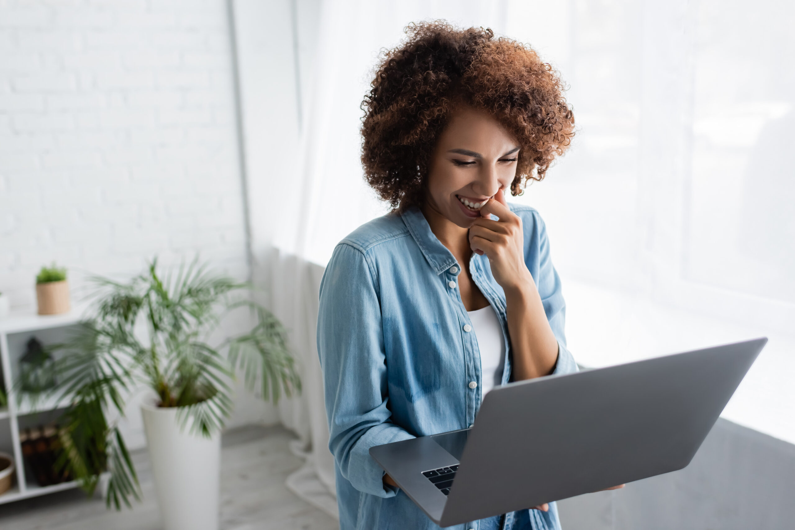 cheerful woman with curly hair holding laptop while working from home