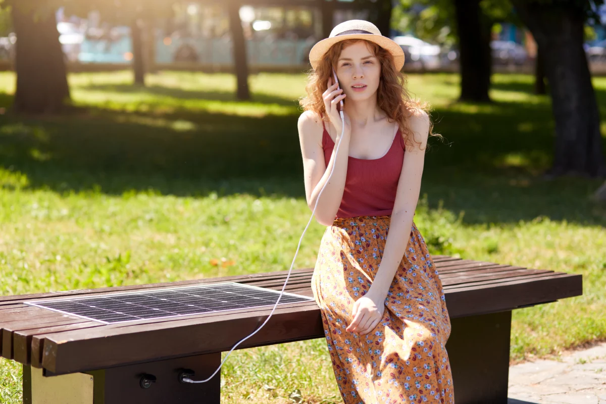 Charming girl charging cell phone via USB on solar panel while talking on the phone outdoor.