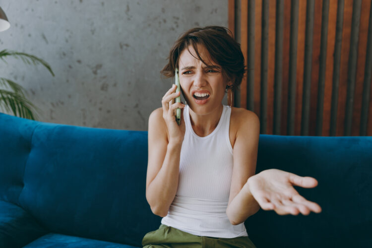 Indignant young woman in white tank shirt spread hands,  while talking on cellphone at home.