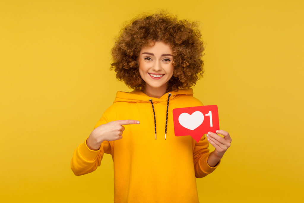 Portrait of joyful curly-haired woman in urban style hoodie pointing at heart like icon, recommending to click on social media button. studio shot isolated on yellow background.