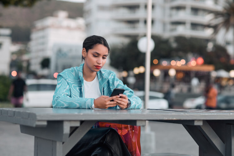 Woman sitting alone, sad at a table outdoor looking at her cell phone.