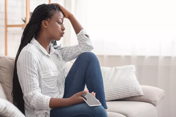 Upset black american woman sitting on the couch, cellphone in hand, thinking about something
