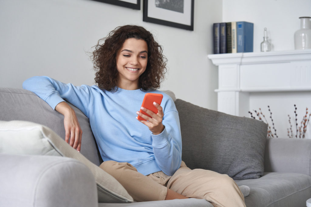 Happy young woman watching video looking at smartphone relaxing on sofa.