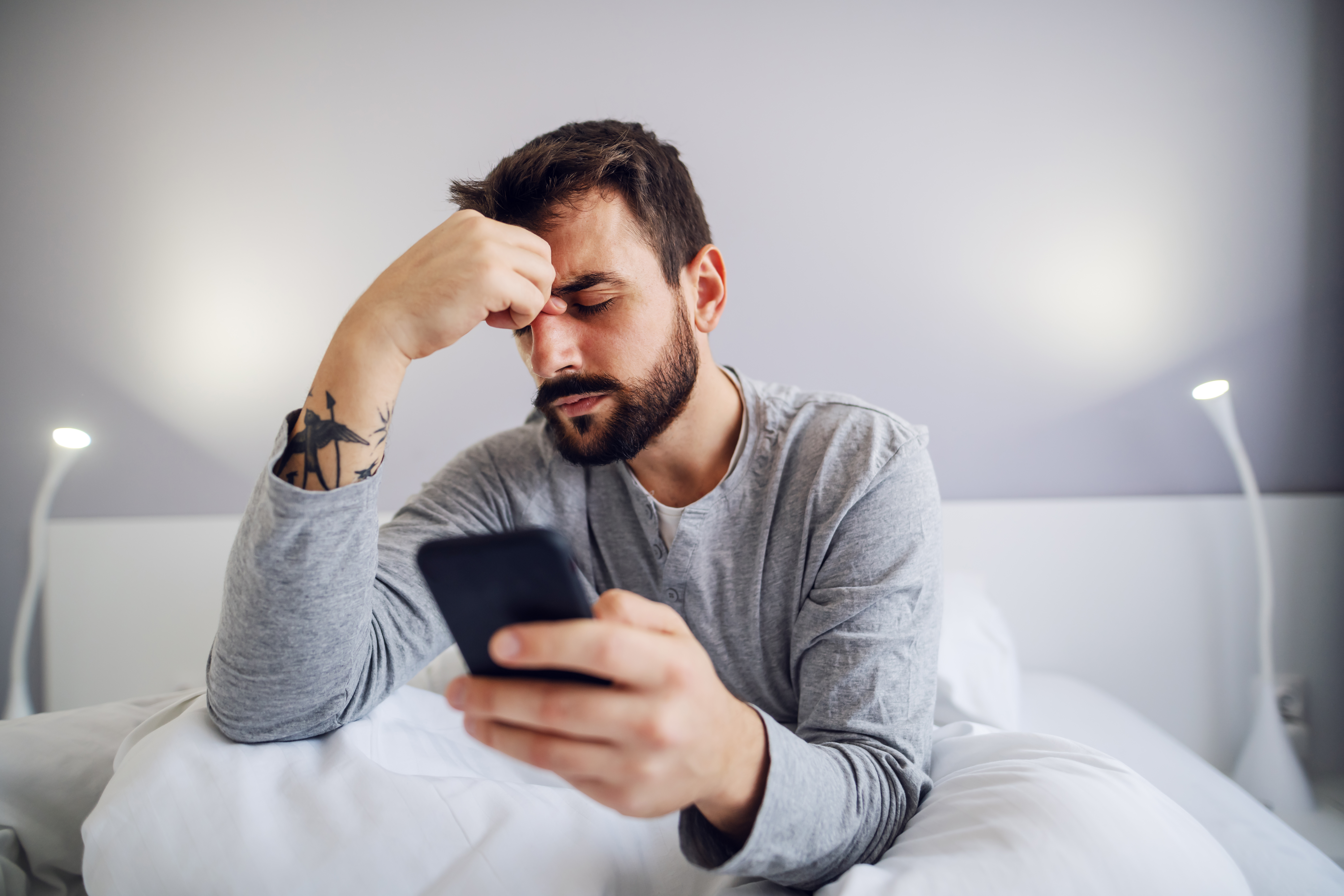Young handsome bearded man sitting in bedroom, holding smart phone