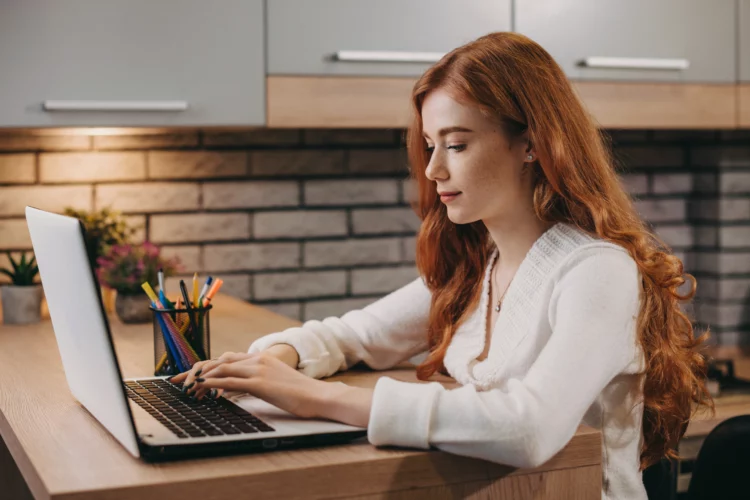 woman with red hair using laptop while sitting at a kitchen table.