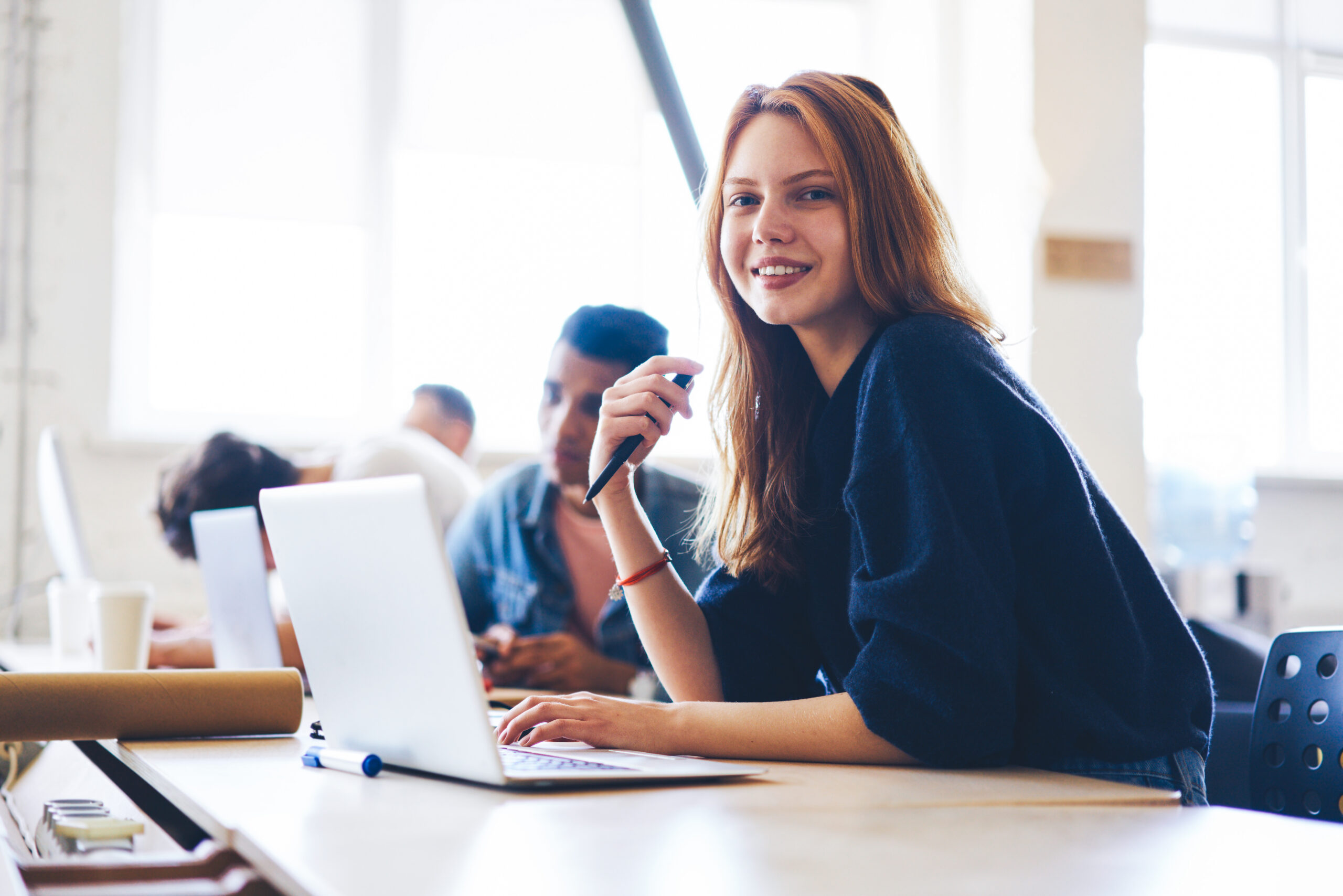 Portrait of charming young female software developer working in modern office with skilled programmers sitting on blurred background,student making researchers doing coursework via laptop in classroom