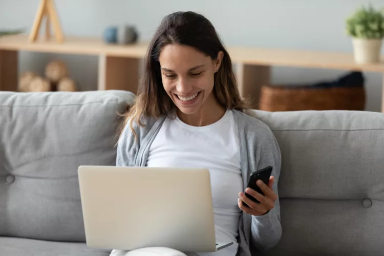 Smiling young lady sitting on sofa at home with computer, making purchases in online store