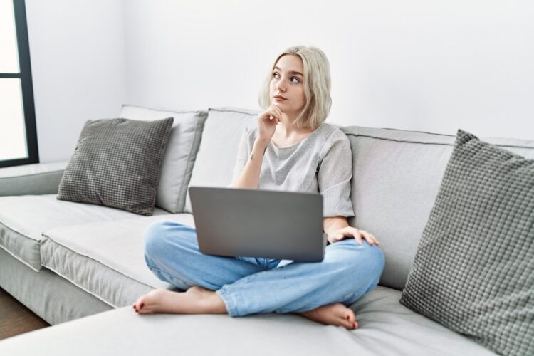 Young woman using laptop at home sitting on the sofa with hand on chin thinking about something.