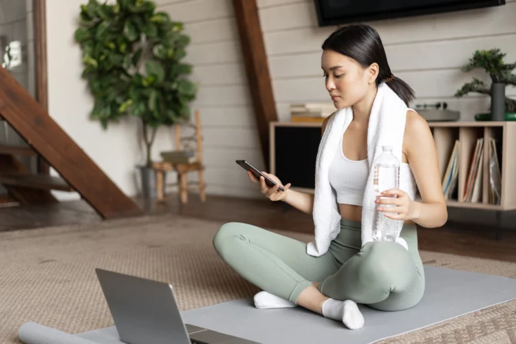 Woman resting from workout at home on floor mat, checking her mobile phone.