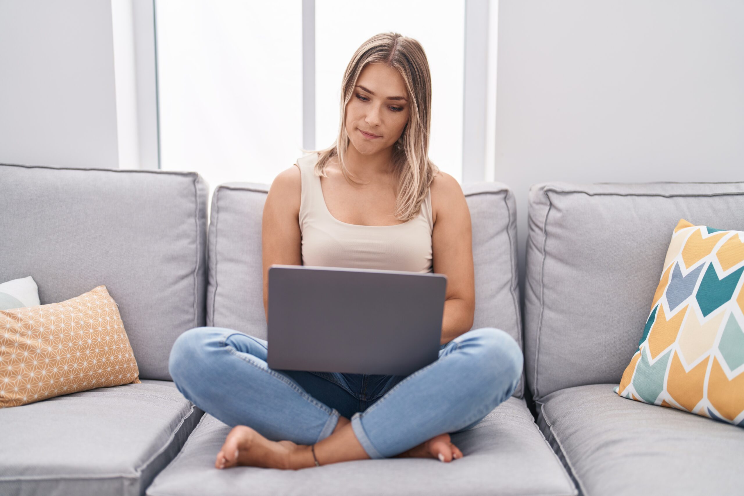 Blonde caucasian woman using laptop at home sitting on the sofa thinking attitude and sober expression looking self confident