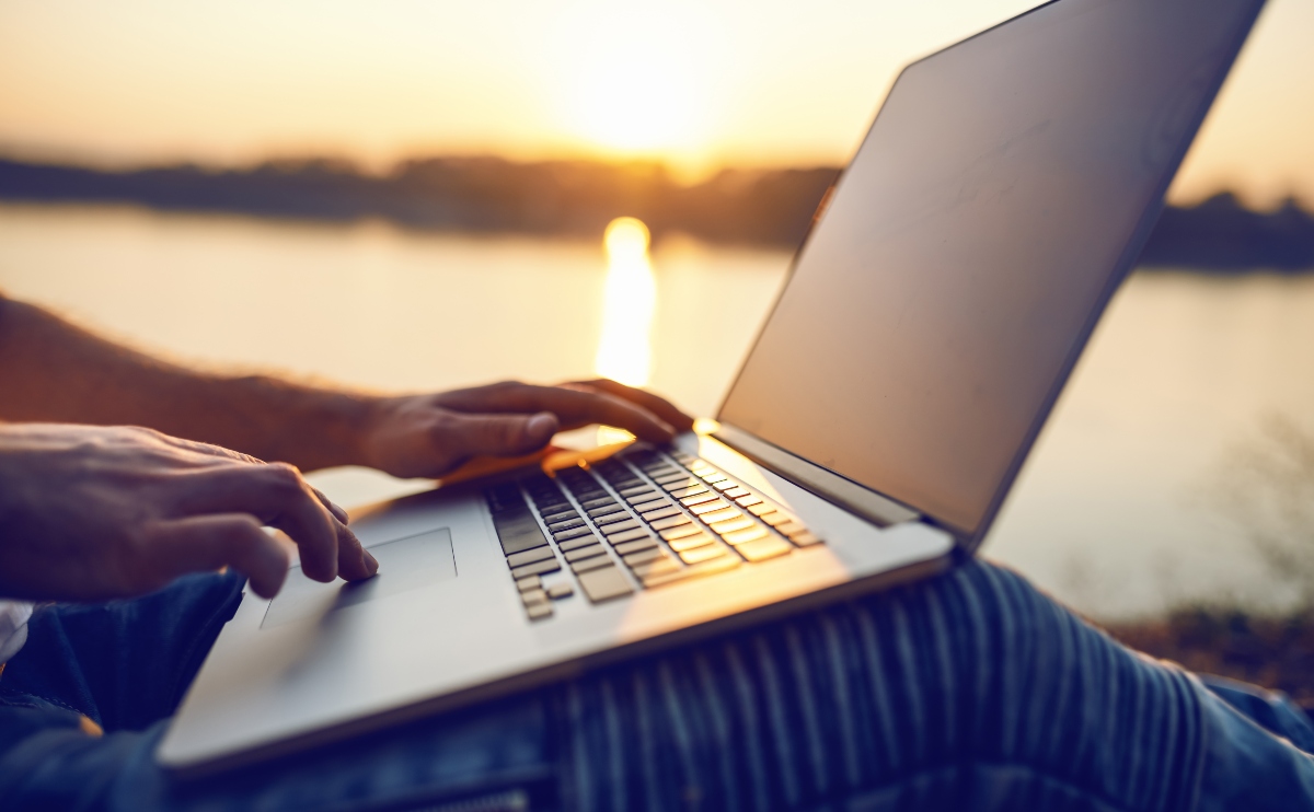 Man sitting in nature and using laptop. In background is river and sunset