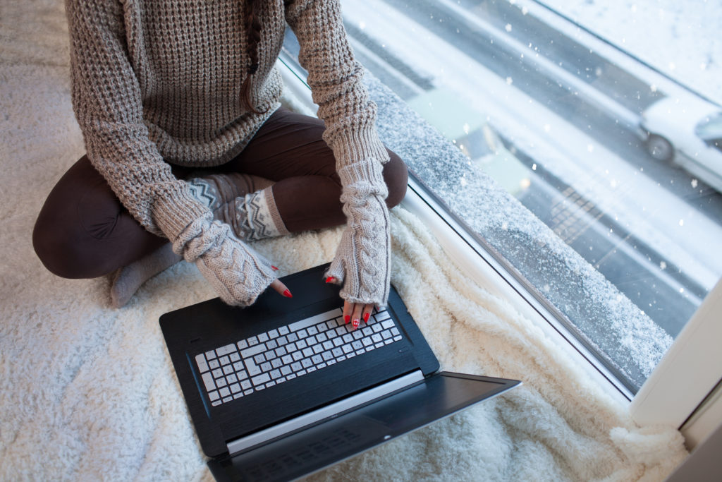 Woman with laptop and gloves sitting at home by the window. 