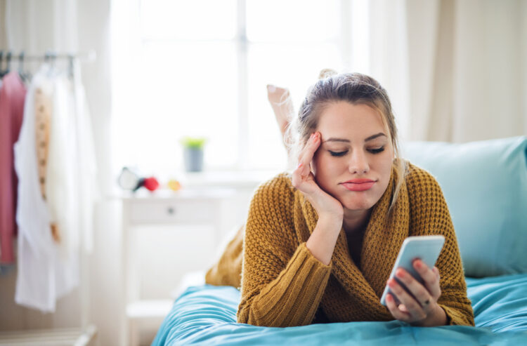 Frustrated young woman with smartphone lying on bed indoors at home.