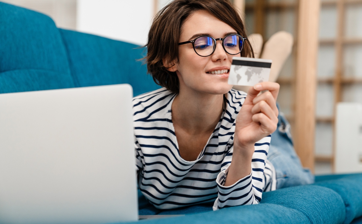 Happy beautiful woman in eyeglasses using credit card and laptop on couch at home