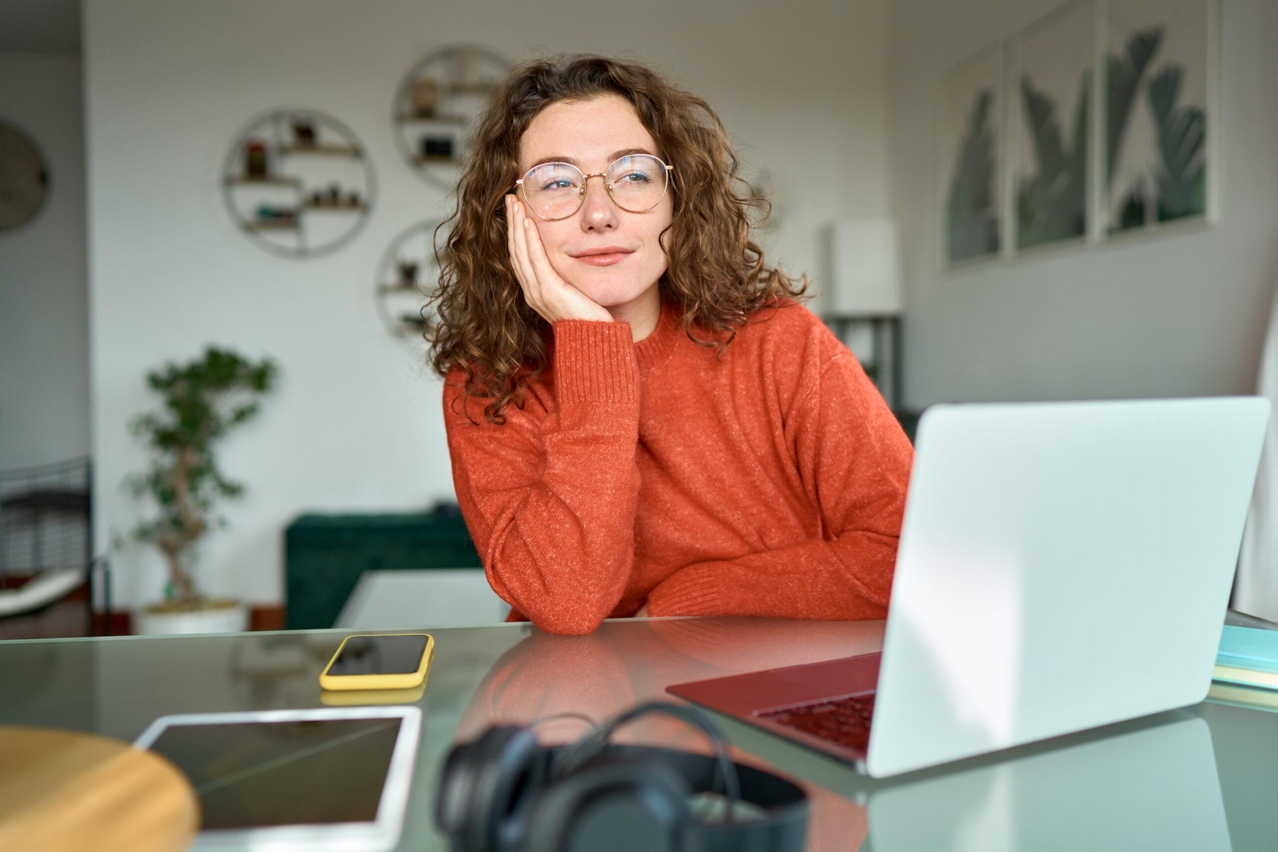 Young woman with glasses and curly hair thinking while working on her laptop at home 