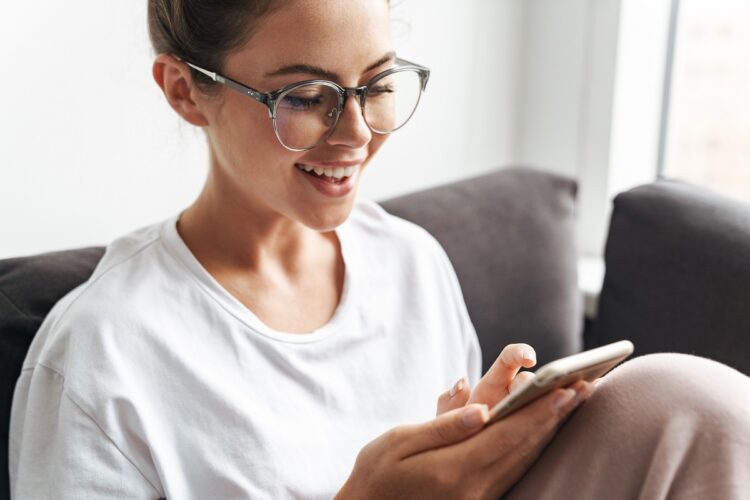 Young woman smiling and typing on cellphone while sitting on sofa.