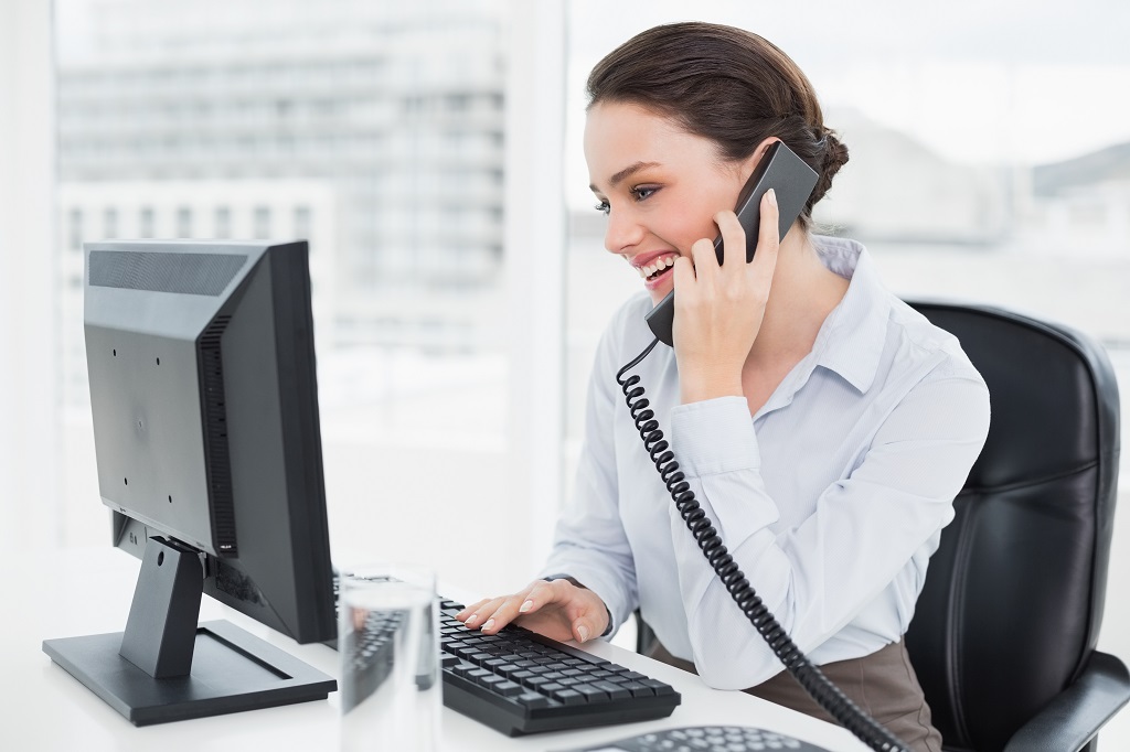 Smiling elegant businesswoman using landline phone and computer in a bright office.