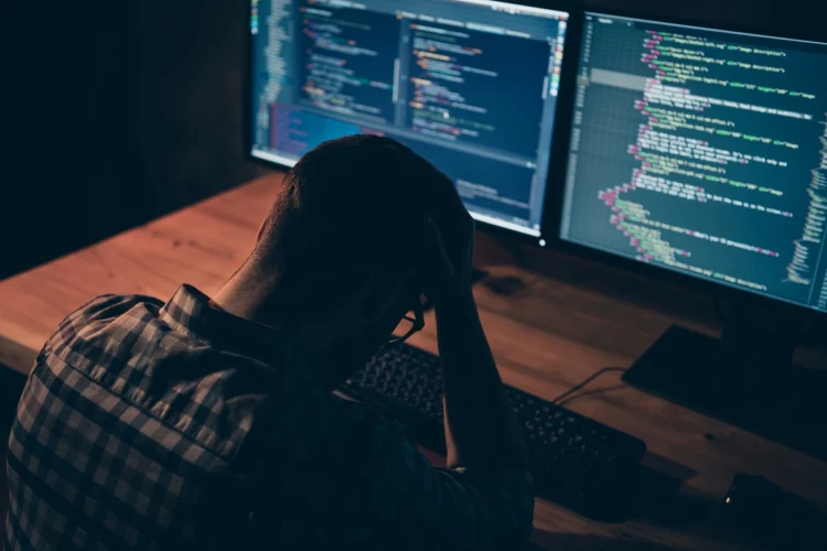 rear of a a frustrated male programmer sitting in front of his computer in a dark office.