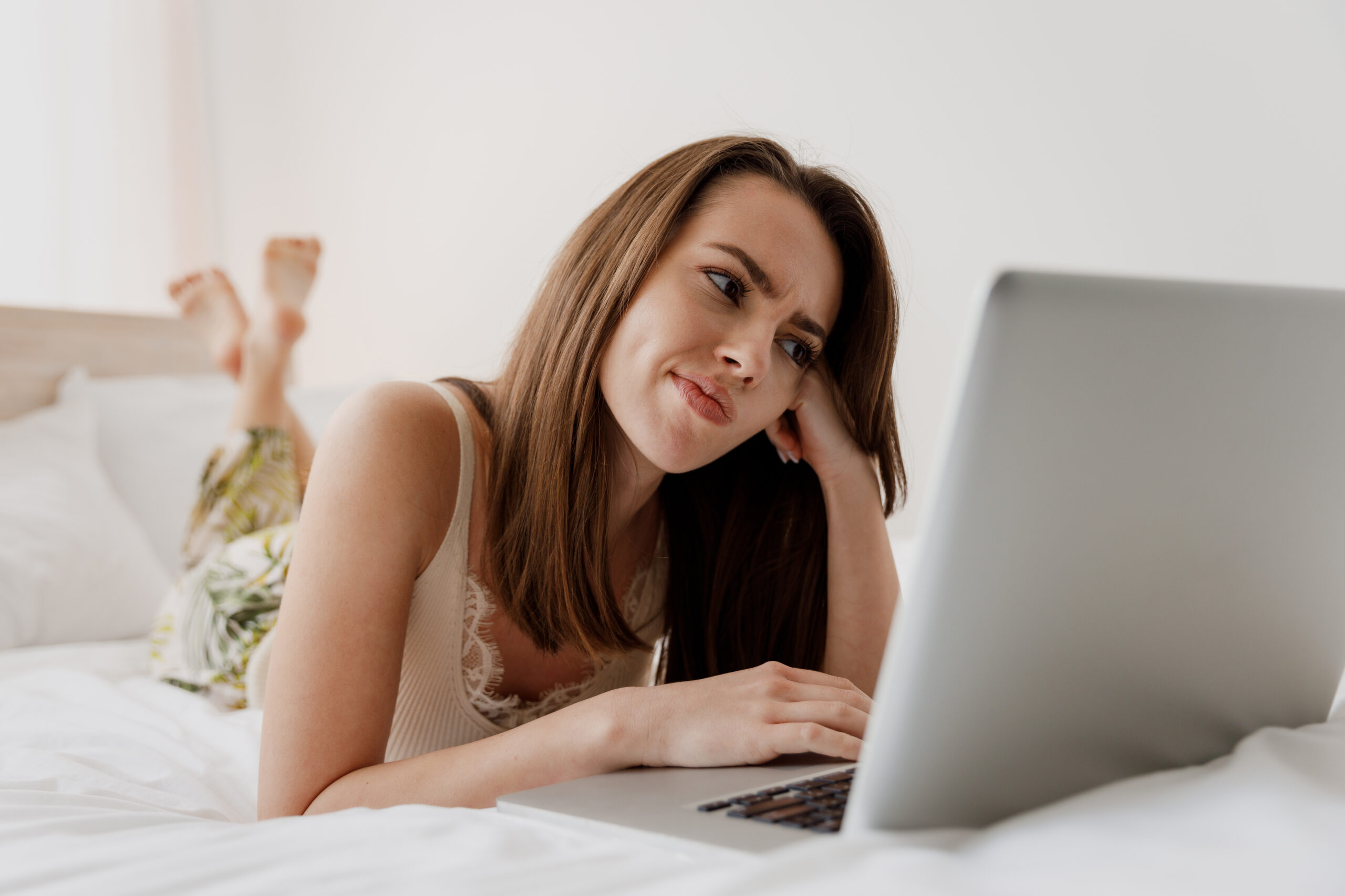 pensive young woman with funny expression lying on her stomach in bed using a laptop