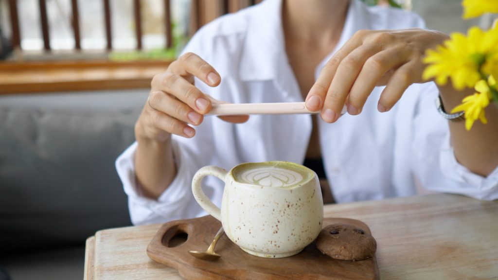 female hands taking a photo of matcha latte served with chocolate chip cookie on a wooden tray.