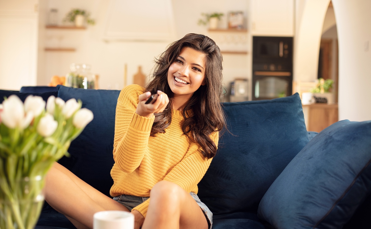 Beautiful smiling woman sitting on a couch at home and watching TV while holding the remote control