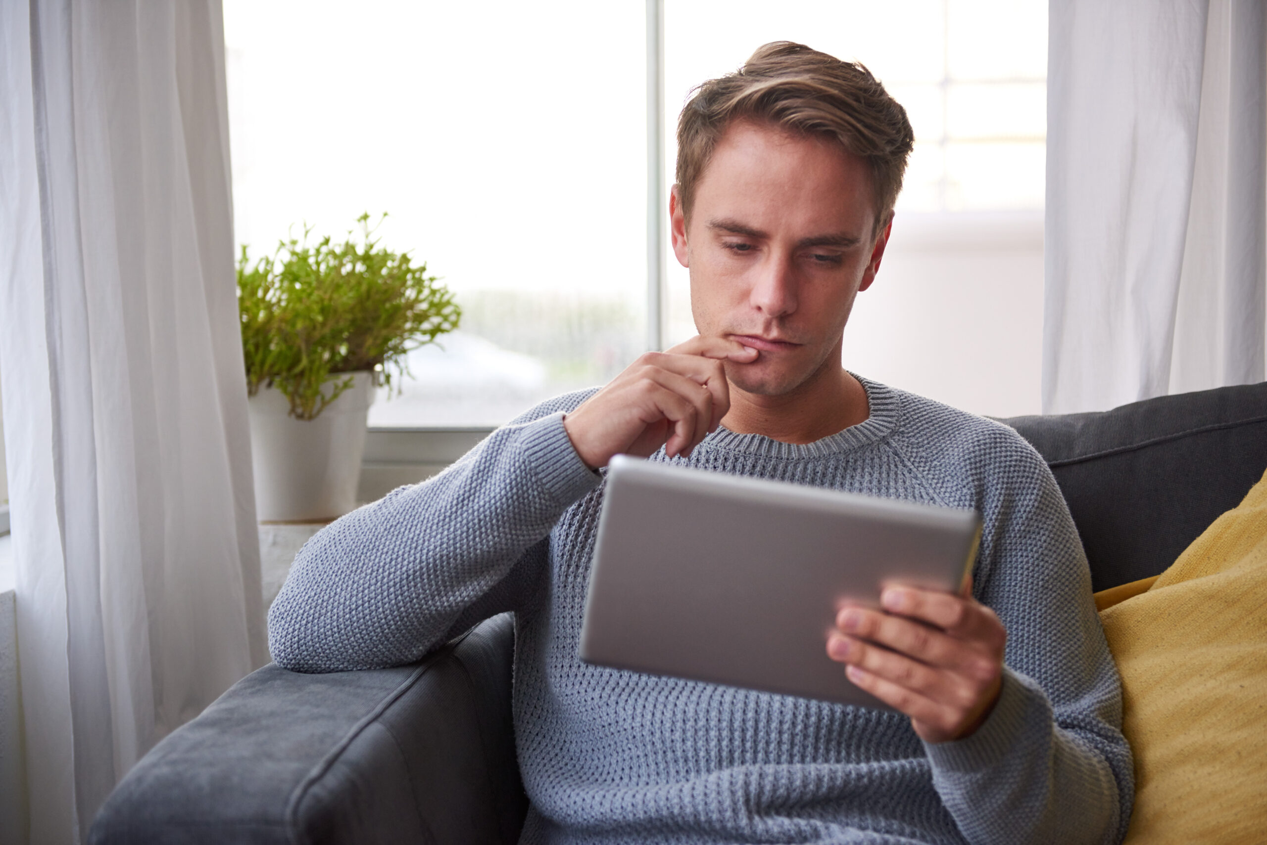 Guy on his couch looking pensive while using a digital tablet