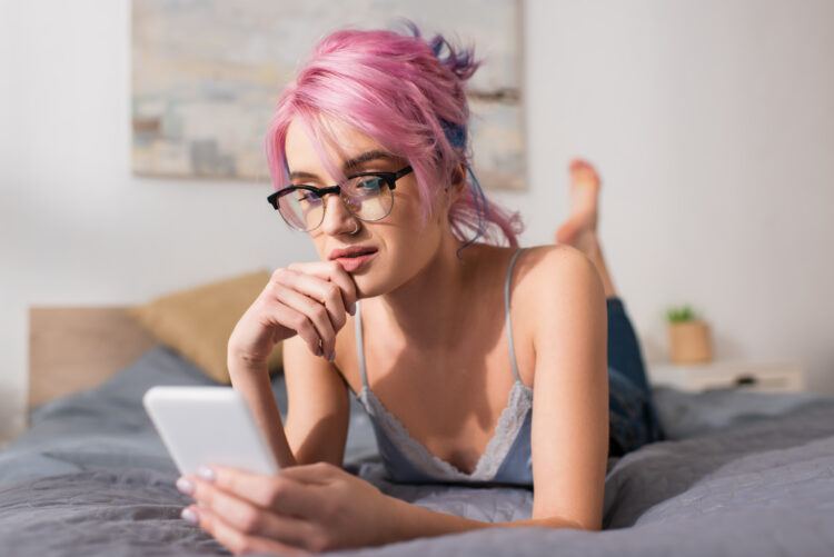 pensive young woman with dyed hair lying on bed while using smartphone.