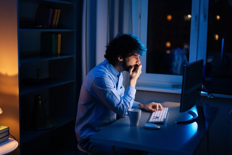 Молодой человек, который выглядит стресс, работая на компьютере в темном домашнем офисе ночью