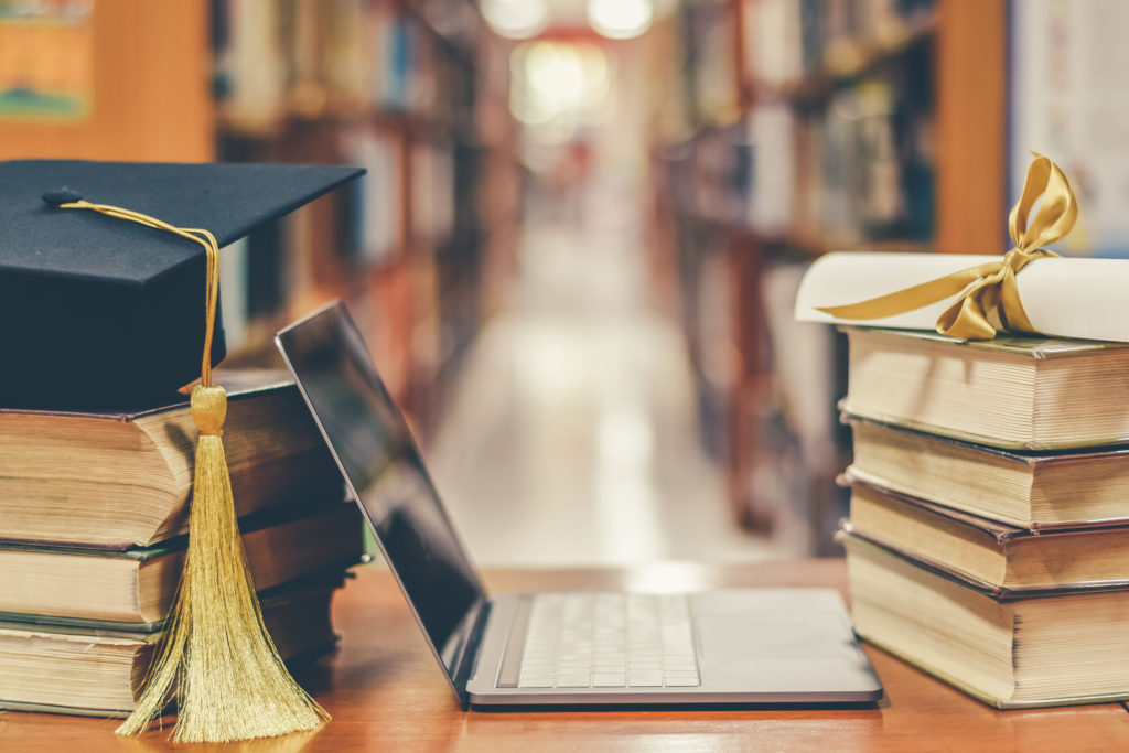 E-learning class and internet online education success with IT computer laptop, graduation hat, academic cap, mortarboard and degree certificate on books in class or library study room.