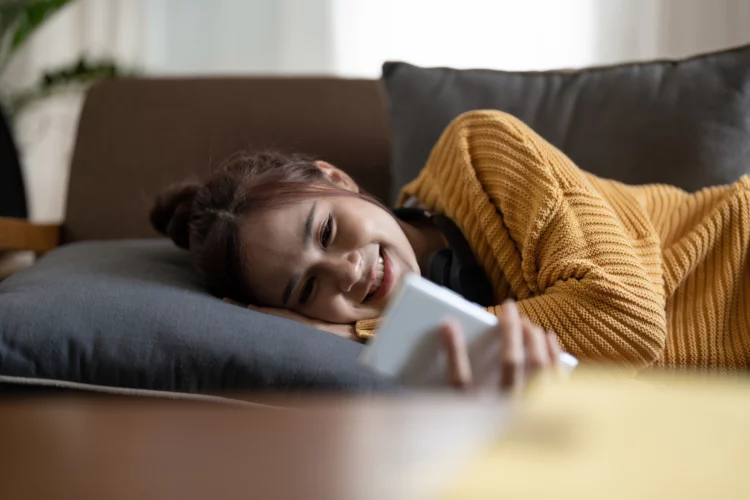 Happy young asian woman lying on sofa at living room and using mobile phone at home