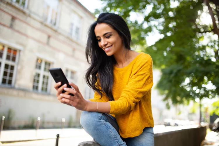 smiling young beautiful woman sitting outdoor, using her smartphone.