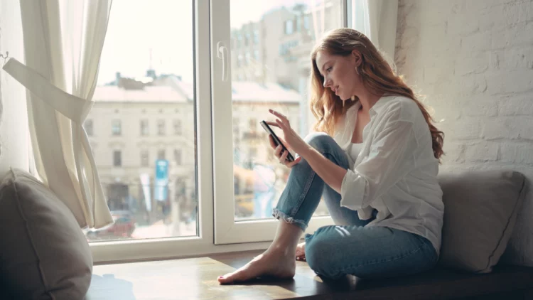 Charming young woman sitting on a windowsill at home and texting on her phone communication