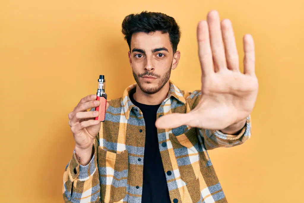 Man holding an e-cigarette while doing a 'stop' sign with his hand, yellow background.