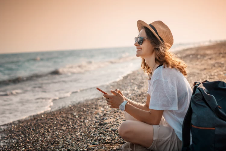 Pretty woman in a hat and sunglasses sitting with a backpack on the seashore at sunset