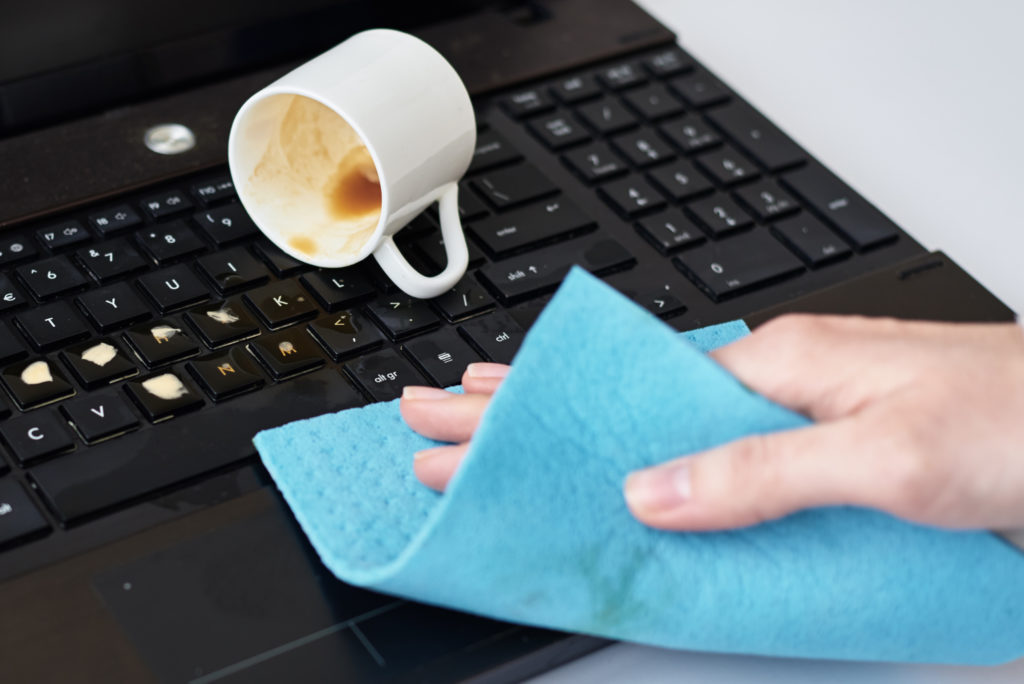  Female hand cleans spilled coffee on laptop keyboard with a blue microfiber cloth.
