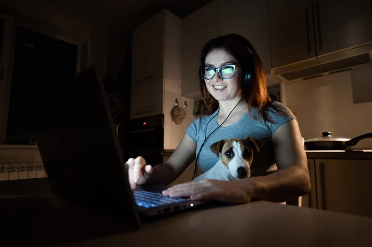 A smiling woman in headphones sits in the dark watching a movie on her laptop in the kitchen with her pet Jack Russell Terrier on her lap.